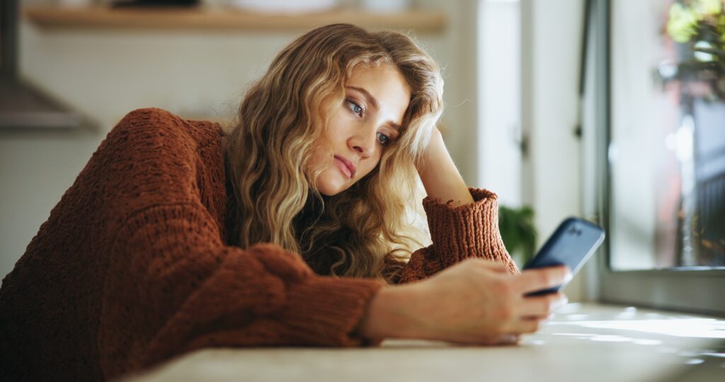 Junge Frau blickt unzufrieden auf ihr Smartphone, unter Druck durch Social-Media-Schönheitsideale.