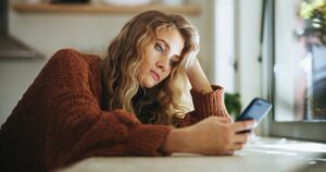 Junge Frau blickt unzufrieden auf ihr Smartphone, unter Druck durch Social-Media-Schönheitsideale.